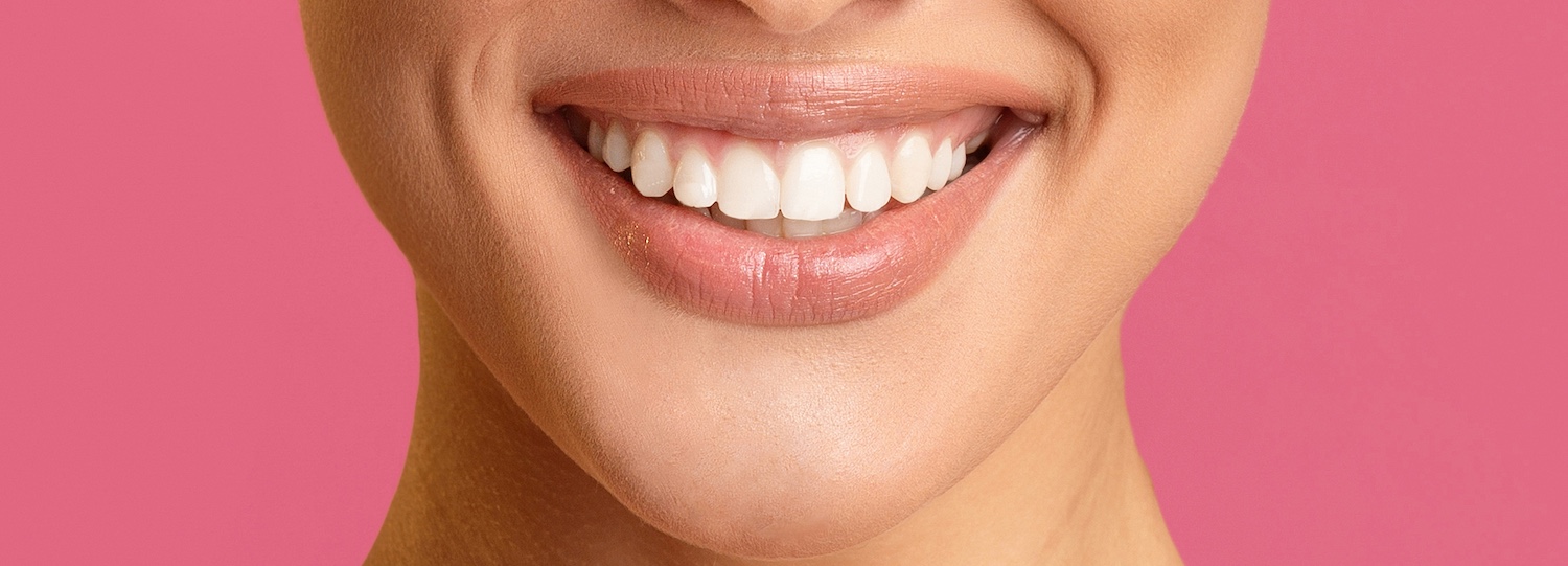 A close-up view of a woman's mouth smiling with her straight white teeth showing in a solid pink background.